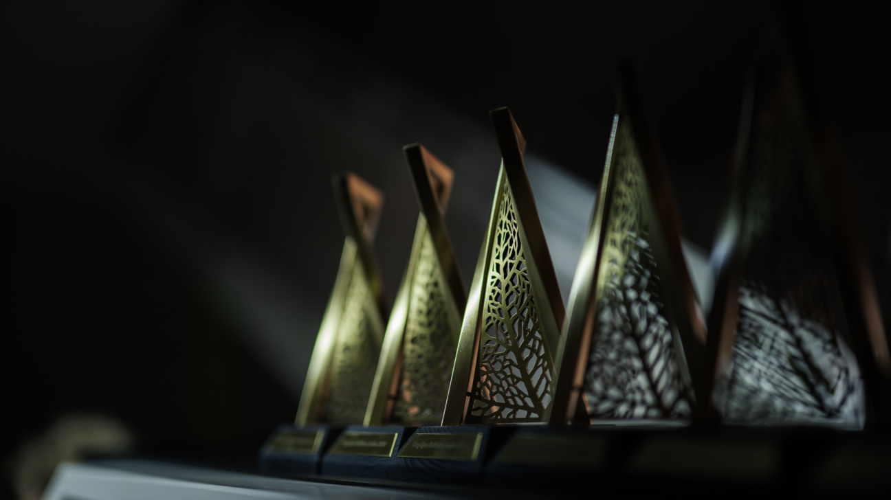 Rindā izkārtoti četri zelta krāsas, trīsstūrveida apbalvojumi ar smalkiem izgrieztiem lapu rakstiem, kas novietoti uz tumša plaukta vājā apgaismojumā. Uz katras pamatnes ir teksts, kas nav skaidri salasāms.