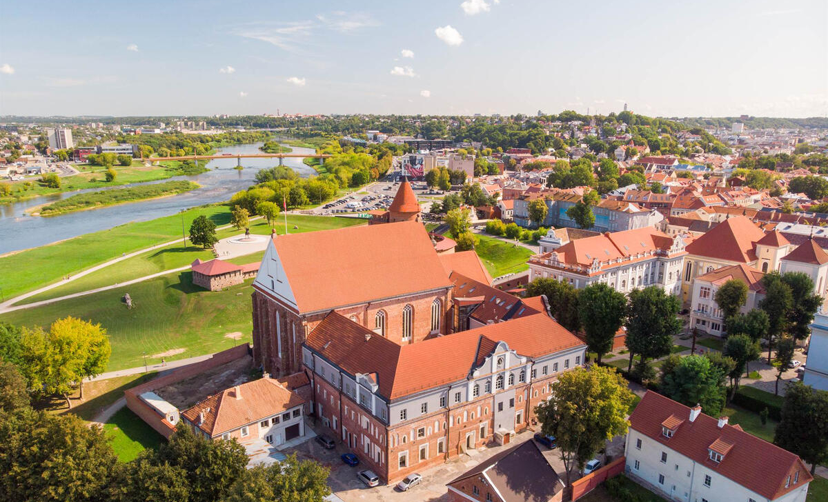 Panorāmas skats uz Kaunas pilsētu ar Kaunas pili, tās sarkanajiem jumtiem upes krastā, ko ieskauj zaļi laukumi un blīvi apbūvēta vecpilsēta saulainā dienā.