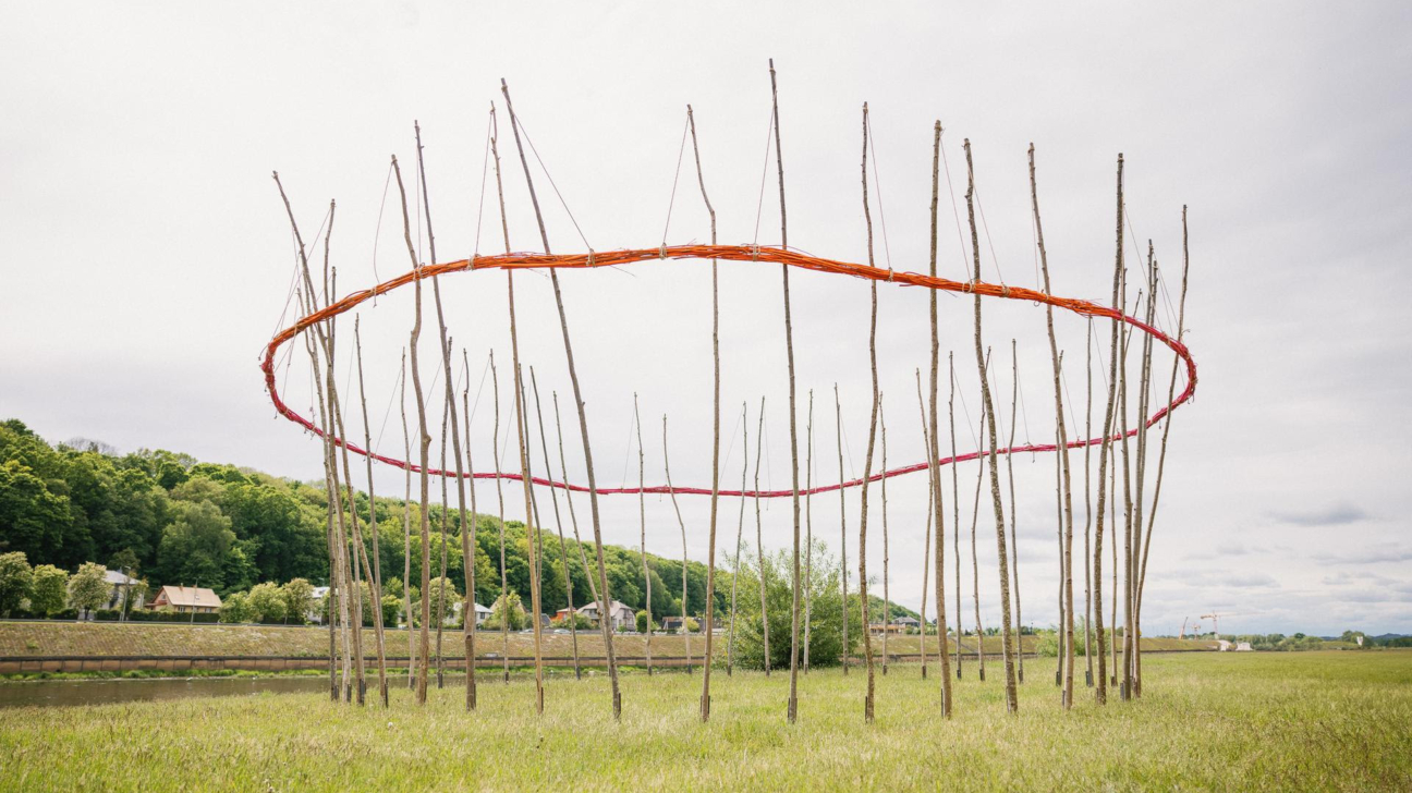 Mākslas instalācija zaļā laukā ar daudziem vertikāliem koka mietiem. Augšpusē starp mietiem pīta oranžsarkana ovāla virkne, zem tās – plānāka sarkana. Aiz instalācijas redzama upe, apdzīvota vieta un mežains kalns zem gaišām, mākoņainām debesīm.