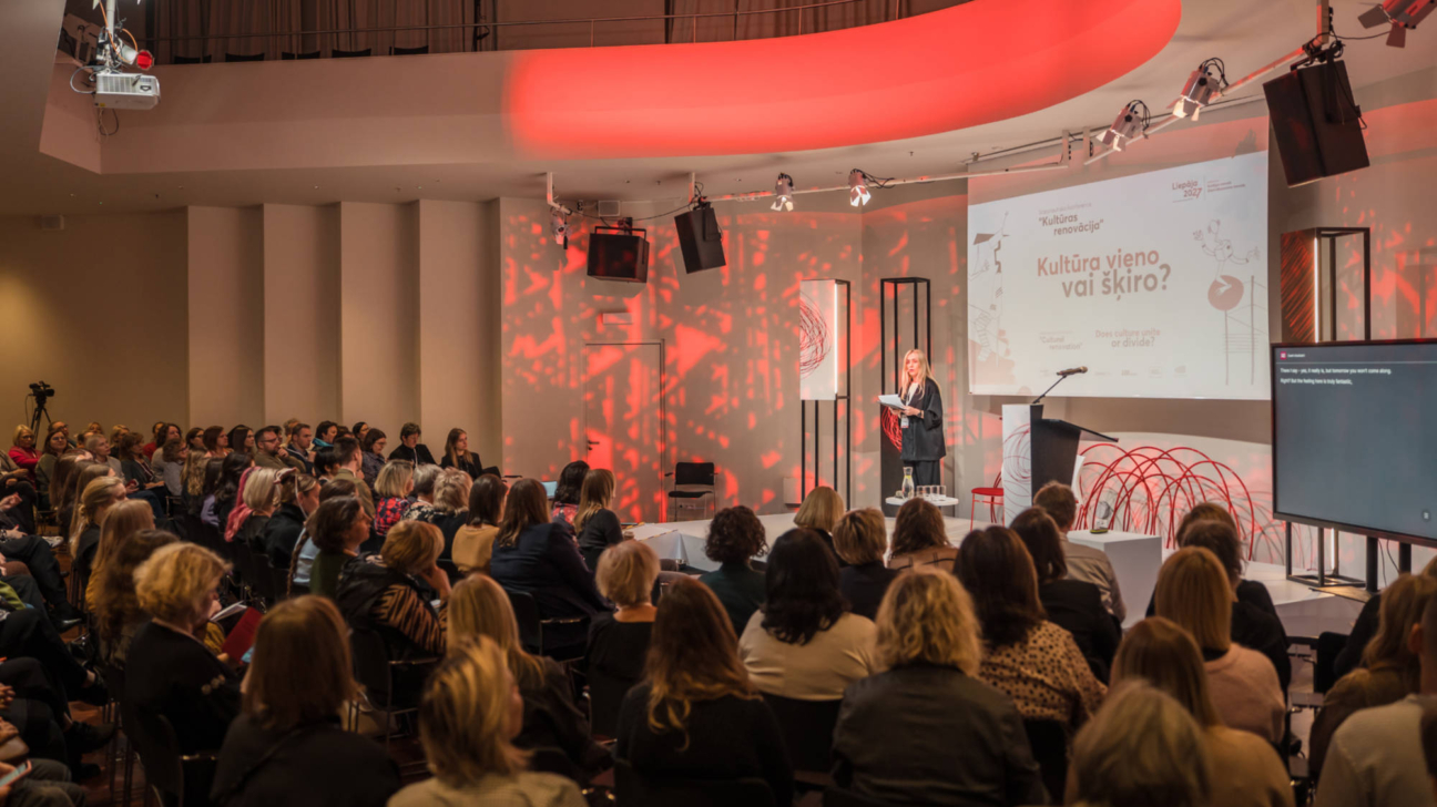 Lektore uzstājas pie pults priekšā lielai auditorijai, kas sēž krēslos un klausās. Aiz viņas esošajā ekrānā redzams uzraksts "Liepāja 2023", "Kultūras renovācija" un jautājums "Kultūra vieno vai šķiro?". Skatuve ir izgaismota sarkanos toņos.