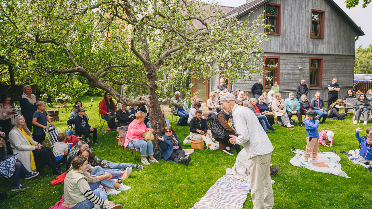 Cilvēki sēž un stāv dārzā pie koka mājas, vērojot priekšnesumu vai pasākumu brīvā dabā; zālienā redzami pieaugušie un bērni.
