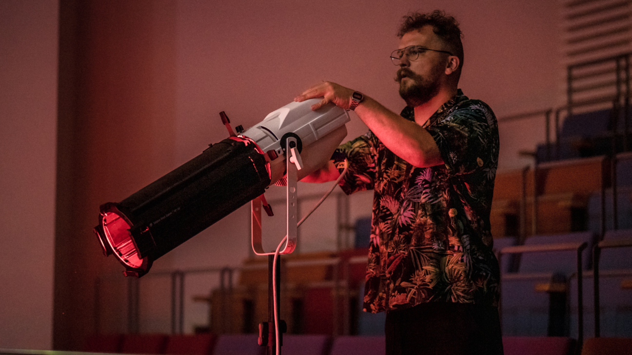 Cilvēks iekštelpās regulē skatuves vai pasākuma prožektoru — rokās tur un pielāgo lielu studijas tipa gaismas lampu uz statīva, telpā redzamas skatītāju sēdvietas.