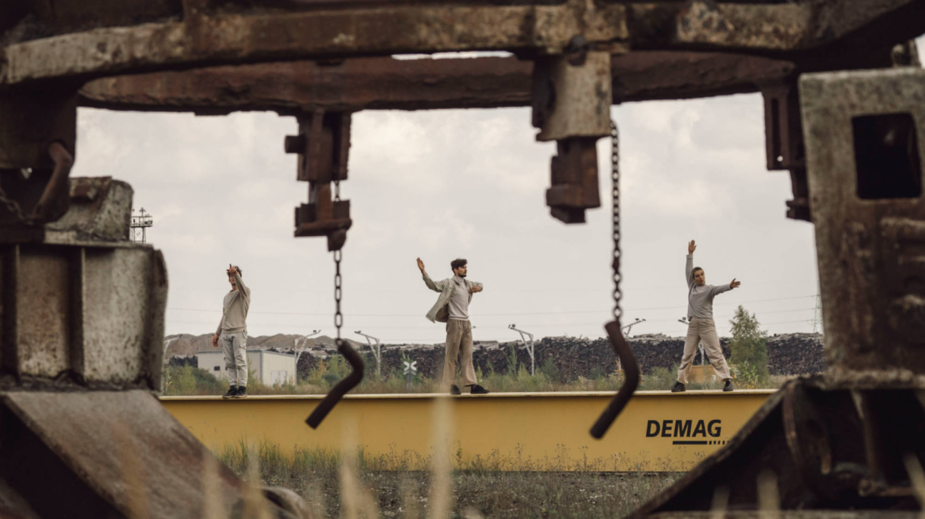 Trīs cilvēki stāv uz dzeltenas sijas industriālā vidē, ko ierāmē rūsainas metāla konstrukcijas. Katram ir paceltas rokas, un uz sijas redzams uzraksts "DEMAG".