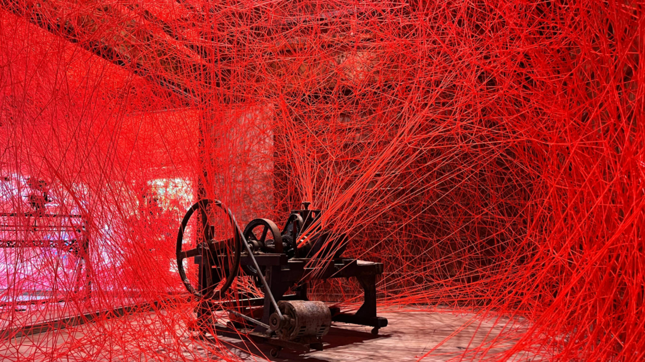 Mākslas instalācija, kurā no vecas, tumšas industriālas mašīnas visā telpā izplūst blīvs, savijies sarkanu diegu tīkls.