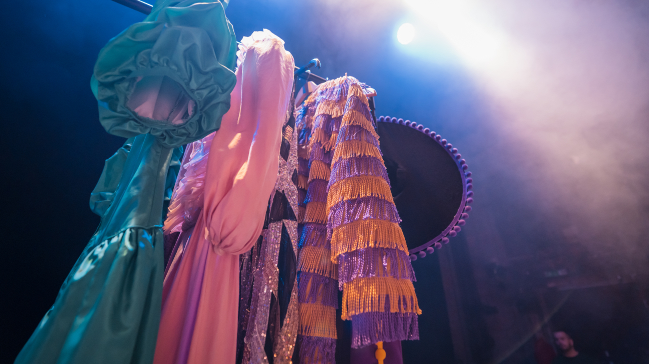 Uz skatuves pakaramā karājas krāsaini teātra kostīmi — zaļa kleita ar kuplām piedurknēm, rozā tērps un dekoratīvs, bārkstains skatuves apģērbs, ko izgaismo prožektoru gaisma.