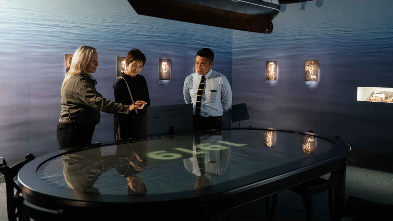 Trīs cilvēki muzejā pēta interaktīvu galdu ar digitālu projekciju; telpā pie sienām redzami vēsturiski portreti un virs galda novietots kuģa modelis