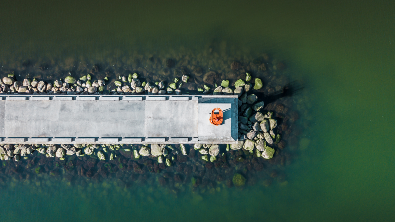 Skats no putna lidojuma uz taisnu betona molu, ko ieskauj akmeņi un zaļgani ūdeņi. Mola galā atrodas oranžs glābšanas riņķis.