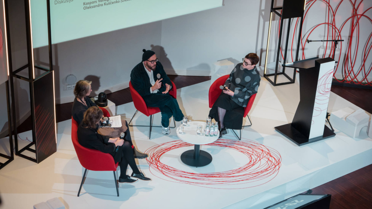 Skats no augšas uz paneļdiskusiju. Četri cilvēki sēž sarkanos krēslos ap baltu galdu.