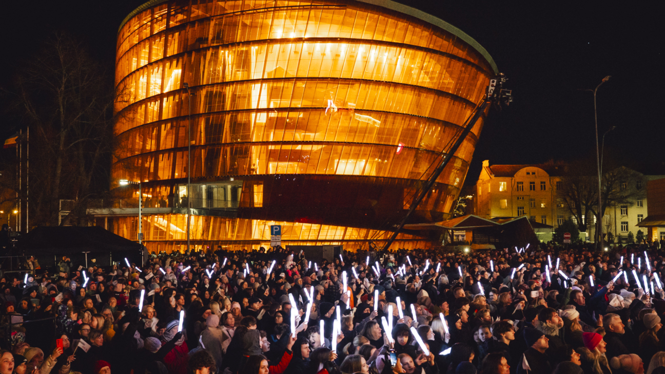 Liels cilvēku pūlis naktī pulcējies pie izgaismotas koncertzāles “Lielais dzintars”, daudzi rokās tur gaismas nūjiņas vai telefonus