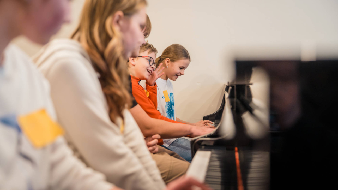 Vairāki bērni sēž rindā un spēlē klavieres. Divi no viņiem, zēns ar brillēm un meitene, smejas un smaida, spēlējot.