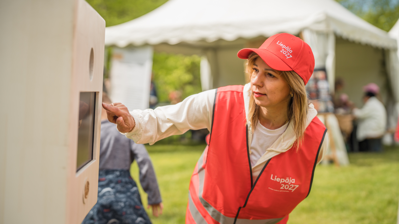 Sieviete ar sarkanu “Liepāja 2027” cepuri un atstarojošu vesti āra pasākumā nospiež interaktīvas ierīces ekrānu; fonā redzama telts un cilvēki.