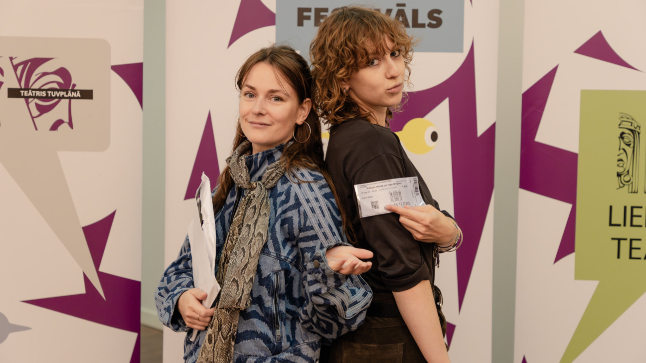 Divas jaunas sievietes, stāvot ar mugurām viena pret otru, pozē kameras priekšā. Kreisā sieviete smaidot tur baltu mapi un rāda ar atvērtu plaukstu. Labā sieviete ar cirtainiem matiem rotaļīgi skatās un tur biļeti ar uzrakstiem "Sieviešu veltes un Suņa fantāzijas", "Liepājas Teātris" un cenu "€14.00". Fonā krāsaini plakāti ar uzrakstiem "FESTIVĀLS" un "TEĀTRIS TUVPLĀNĀ".