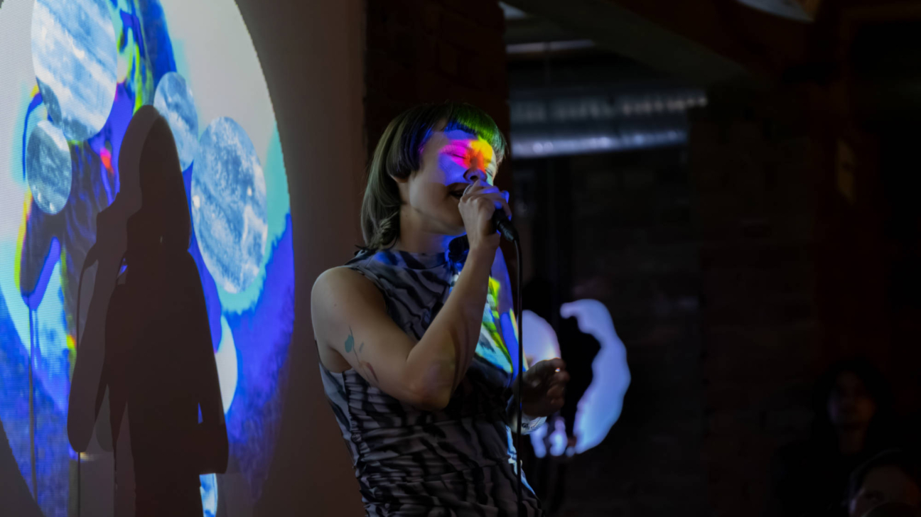 Persona ar īsiem matiem dzied mikrofonā. Varavīksnes krāsu gaisma apgaismo viņas seju. Aizmugurē uz sienas tiek projicētas abstraktas zili baltas formas, kas met dziedātājas ēnu.