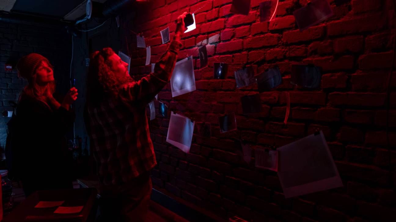 Divi cilvēki tumšā telpā, ko apgaismo sarkana gaisma, pie ķieģeļu sienas, kas noklāta ar virknēs sakārtotām fotogrāfijām. Viens no viņiem karina jaunu attīstītu fotogrāfiju, otrs stāv blakus un vēro.
