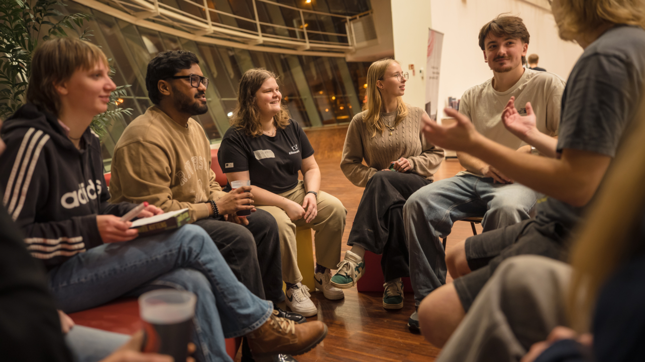 Jauniešu grupa sēž aplī iekštelpās un sarunājas, daļai rokās ir dzērienu glāzes, viens dalībnieks runā, kamēr pārējie klausās
