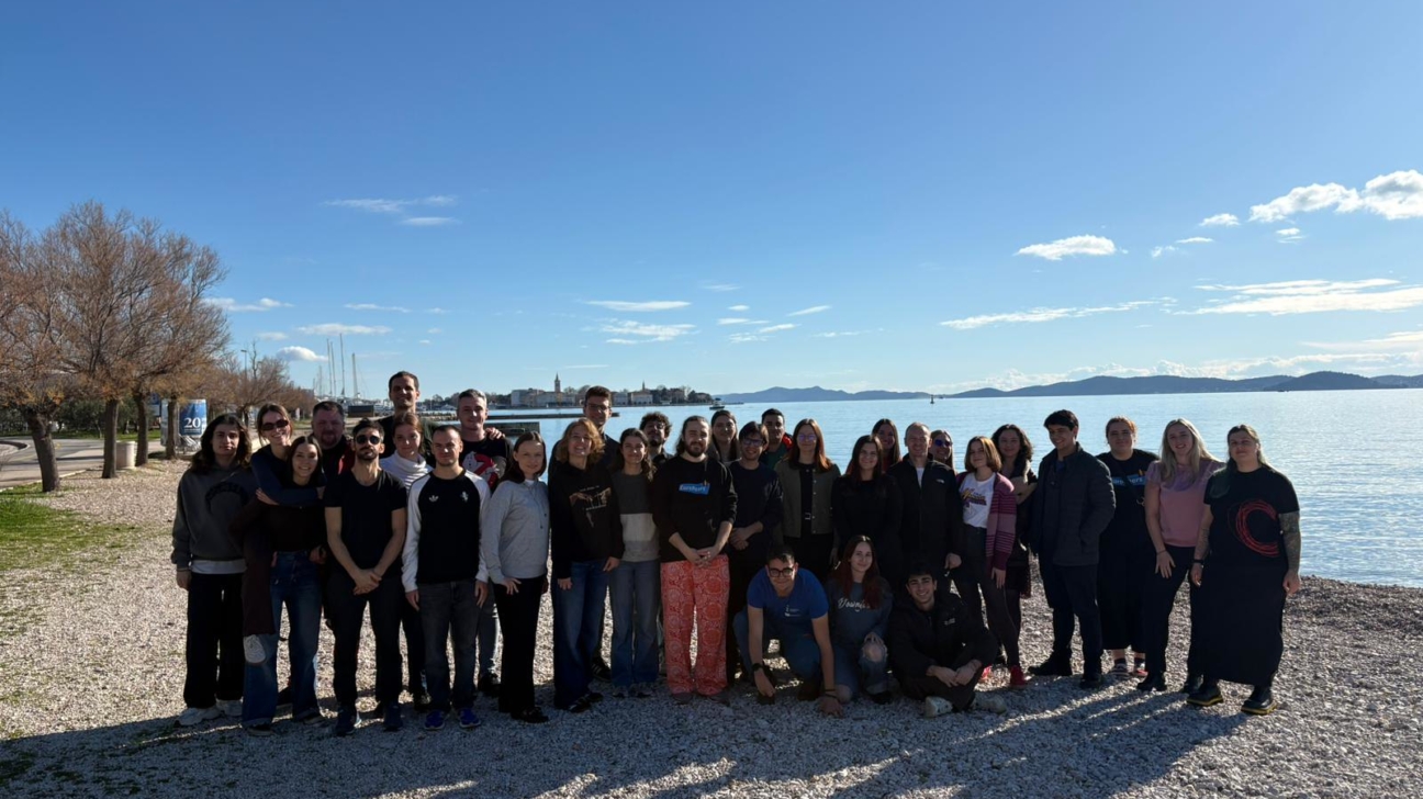 Liela cilvēku grupa pozē fotogrāfijai uz oļu pludmales pie zilas jūras. Fonā redzama pilsētas panorāma un tāls krasts zem skaidrām debesīm.