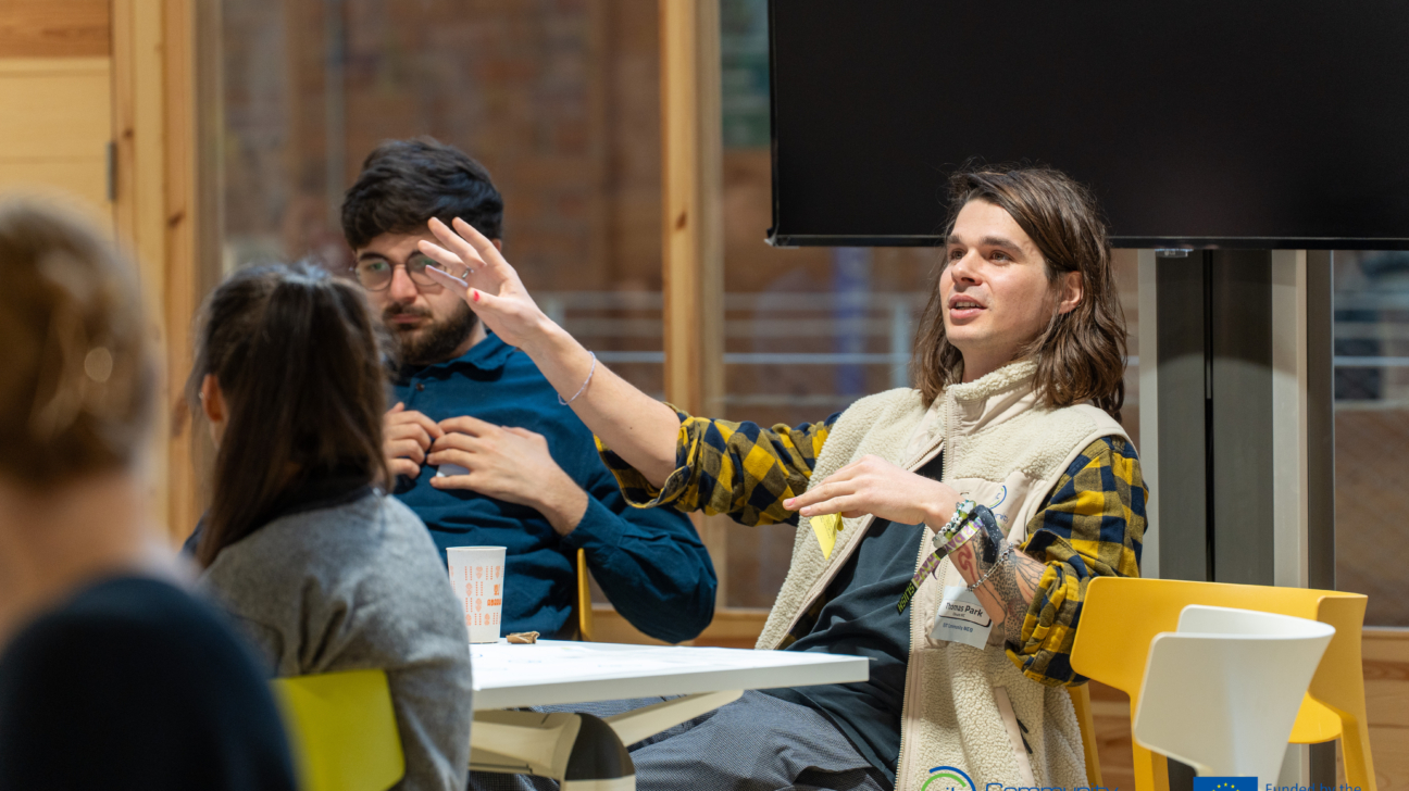 Jauniešu diskusija vai darbnīca telpā – viens dalībnieks sēž pie galda un runājot žestikulē ar rokām, kamēr citi klausās. Uz galda redzama krūze, fonā ekrāns, apakšā redzami “EIT Community” un Eiropas Savienības finansējuma logotipi