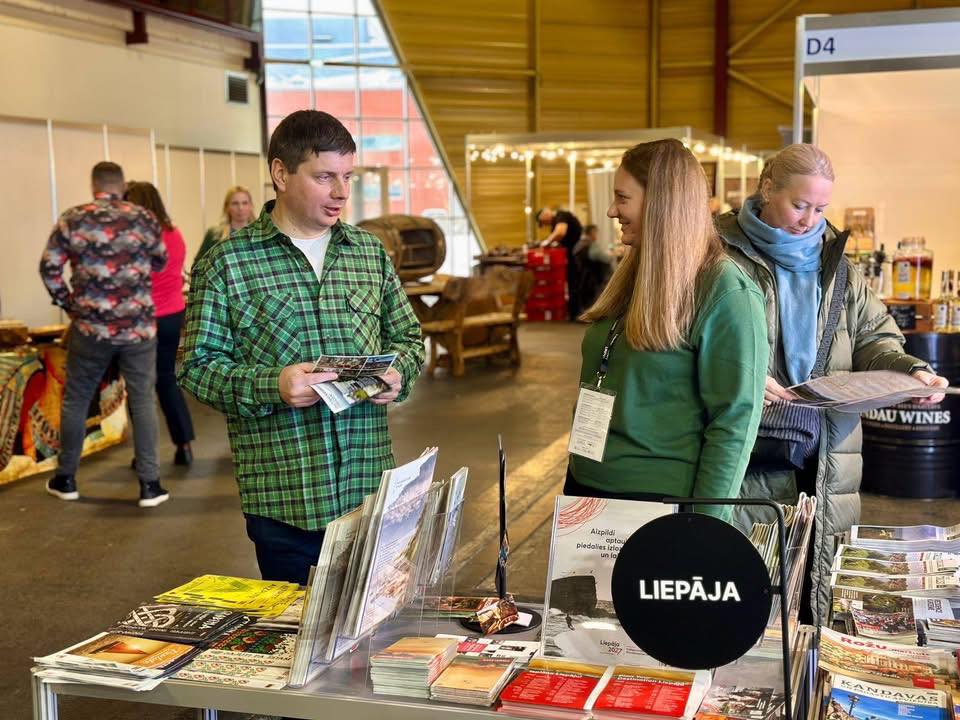 Trīs cilvēki izstādes zālē pie informatīvā stenda. Priekšplānā vīrietis zaļā rūtainā kreklā ar bukletiem rokās runā ar sievieti zaļā džemperī, kura smaida. Blakus sieviete zilā šallē un virsjakā pēta bukletu. Uz galda starp dažādiem bukletiem un žurnāliem ir melna apaļa zīme ar tekstu "LIEPĀJA". Fonā redzami citi stendi, tostarp viens ar mucu, uz kuras rakstīts "DAU WINES".