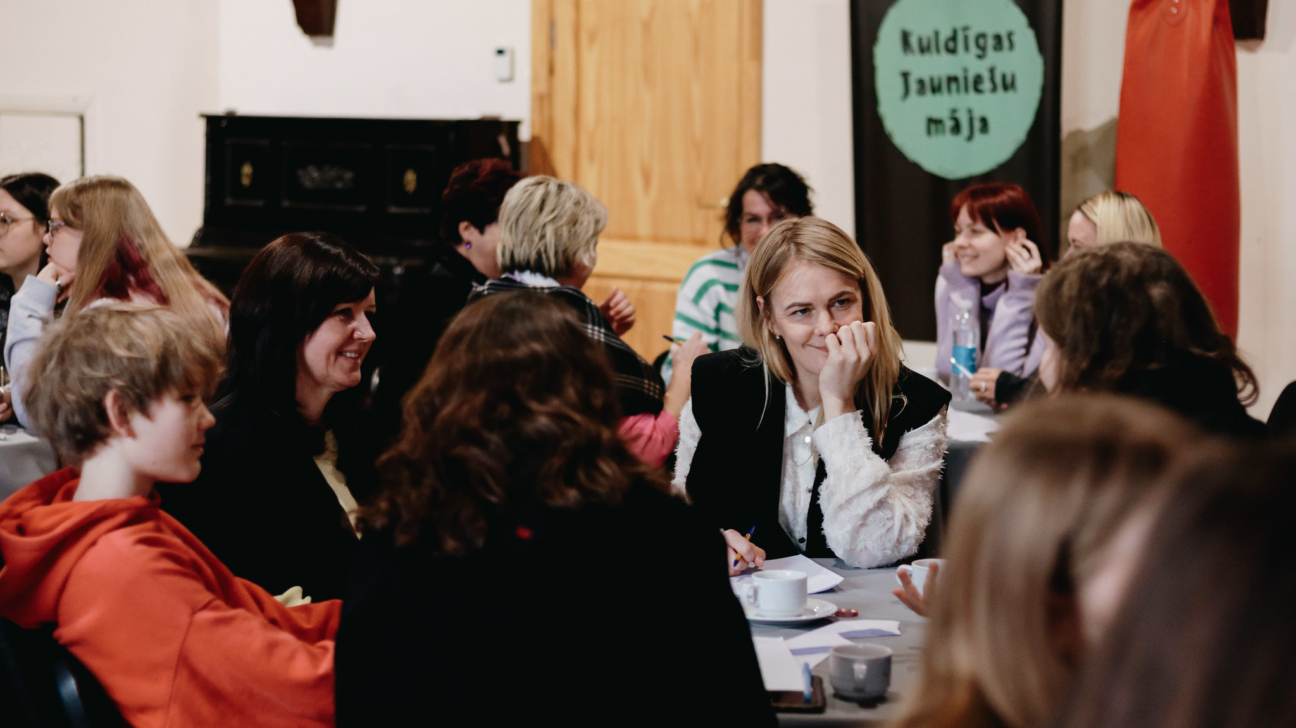 Sanāksme vai pasākums, kurā dažāda vecuma cilvēki sēž pie galdiem, smaida un sarunājas. Priekšplānā gaišmate sieviete ar roku pie zoda, blakus zēns sarkanā džemperī. Fonā plakāts ar tekstu "Kuldīgas Jauniešu māja" un sarkans boksa maiss.