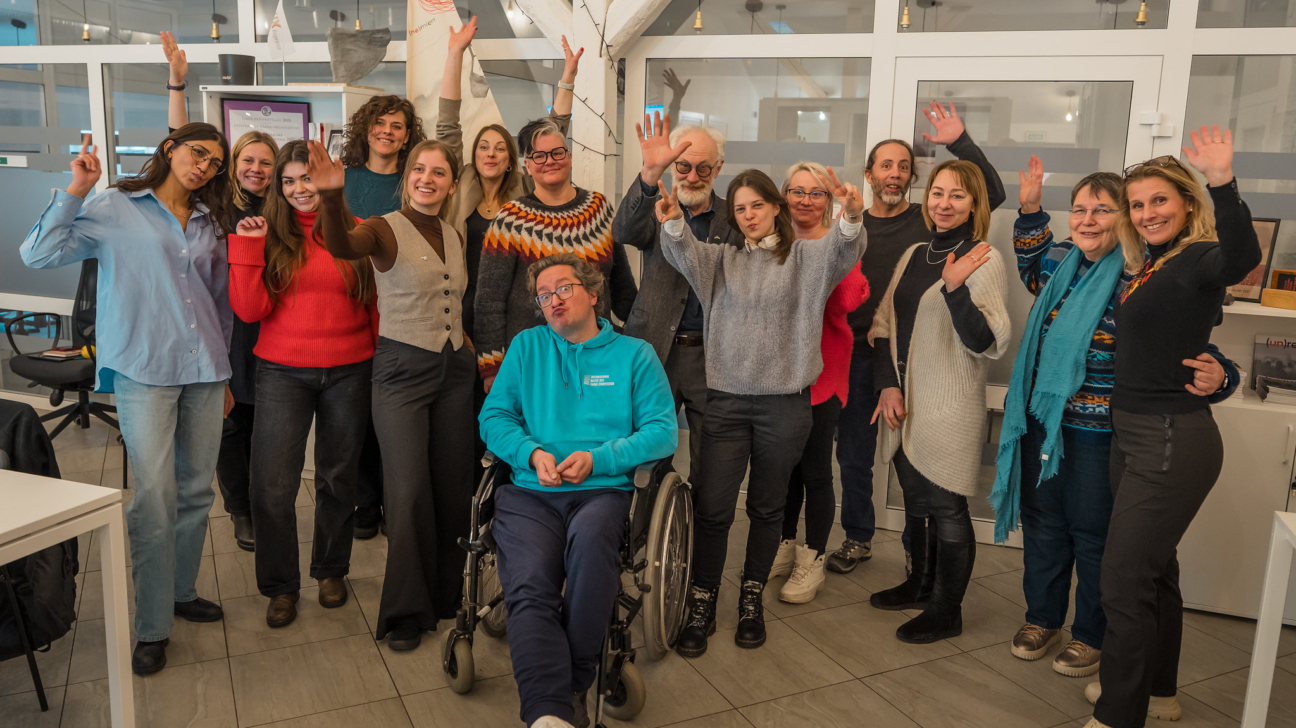 Daudzveidīga aptuveni 20 smaidīgu pieaugušo grupa, no kuriem daži māj ar rokām vai rāda miera zīmes, pozē iekštelpās. Priekšplānā sēž vīrietis ratiņkrēslā, tērpies tirkīza džemperī ar uzrakstu "LATVIJAS KULTŪRAS AKADĒMIJA", ar rotaļīgu sejas izteiksmi.