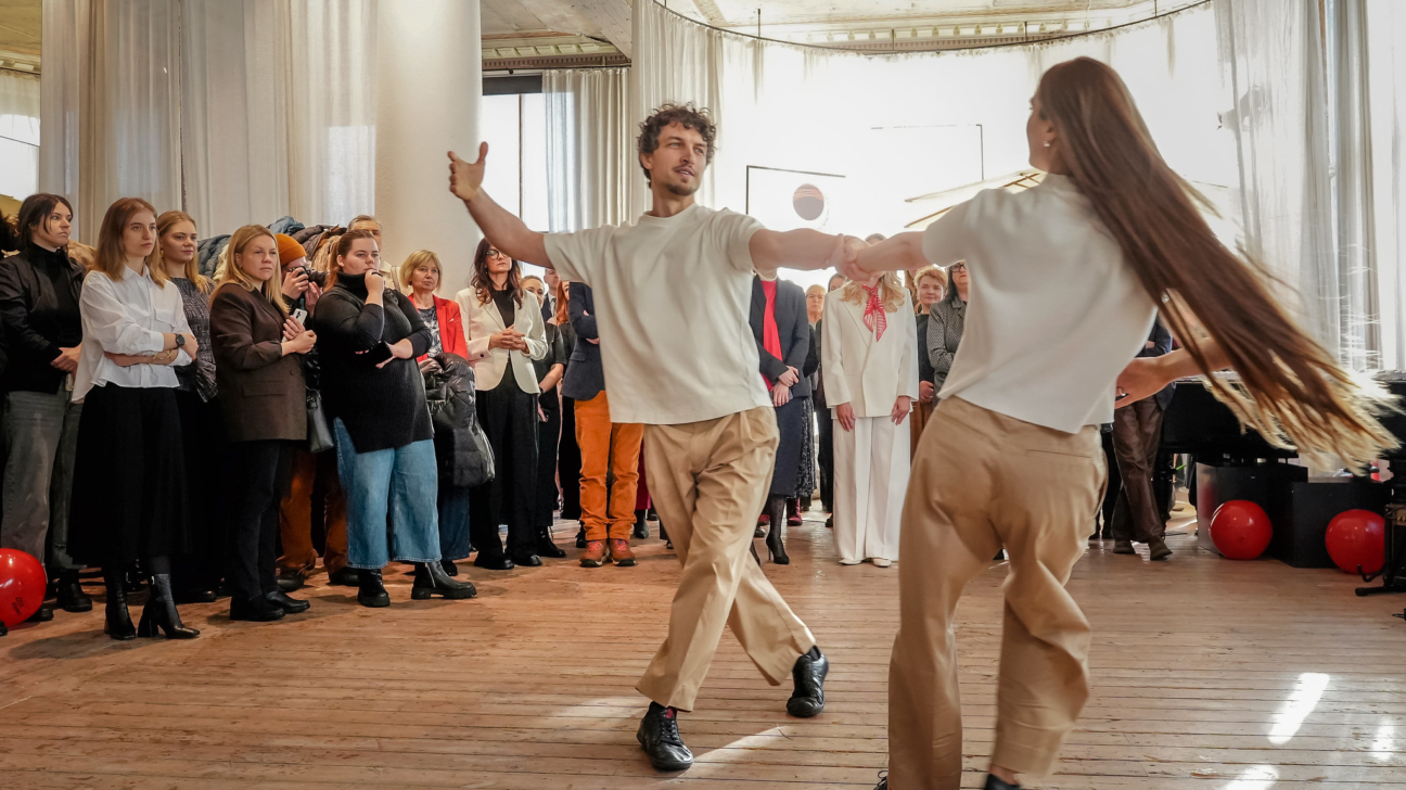 Vīrietis un sieviete dejo uz koka grīdas, turoties rokās, kamēr viņus vēro skatītāju pūlis gaišā telpā. Sievietes garie mati plīvo kustībā.