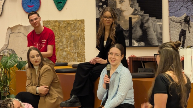 Grupa jaunu cilvēku sēž uz oranžiem pakāpieniem, sarunājoties. Viena sieviete tura mikrofonu un smaida. Vīrietim sarkanā T-kreklā ir uzraksts 