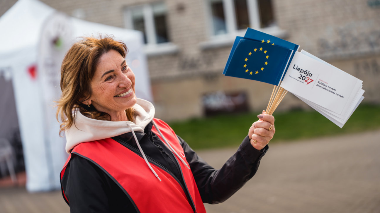 Smaidoša sieviete sarkanā vestē tur rokās vairākus ES karodziņus un baltas kartītes ar tekstu "Liepāja 2027 Eiropas kultūras galvaspilsēta Kuldīgas novads Dienvidkurzemes novads".