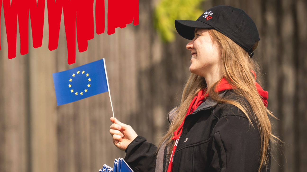 Smaidoša sieviete melnā cepurē ar uzrakstu "Liepāja 2027" un sarkanā džemperī, tur rokā mazu Eiropas Savienības karogu.