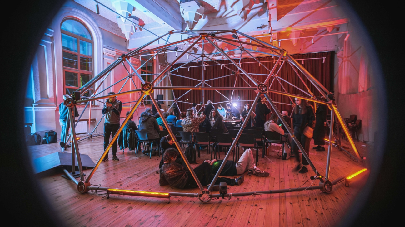 Lielā telpā ar koka grīdu, izgaismotā zilos un oranžsarkanos toņos, centrā atrodas metāliska ģeodēziska kupola konstrukcija ar iestrādātiem oranžiem gaismas elementiem. Apmeklētāji pulcējušies ap kupolu un tā iekšpusē, daži sēž krēslos, daži stāv, bet daži atrodas guļus uz grīdas, vērojot notiekošo pasākumu.