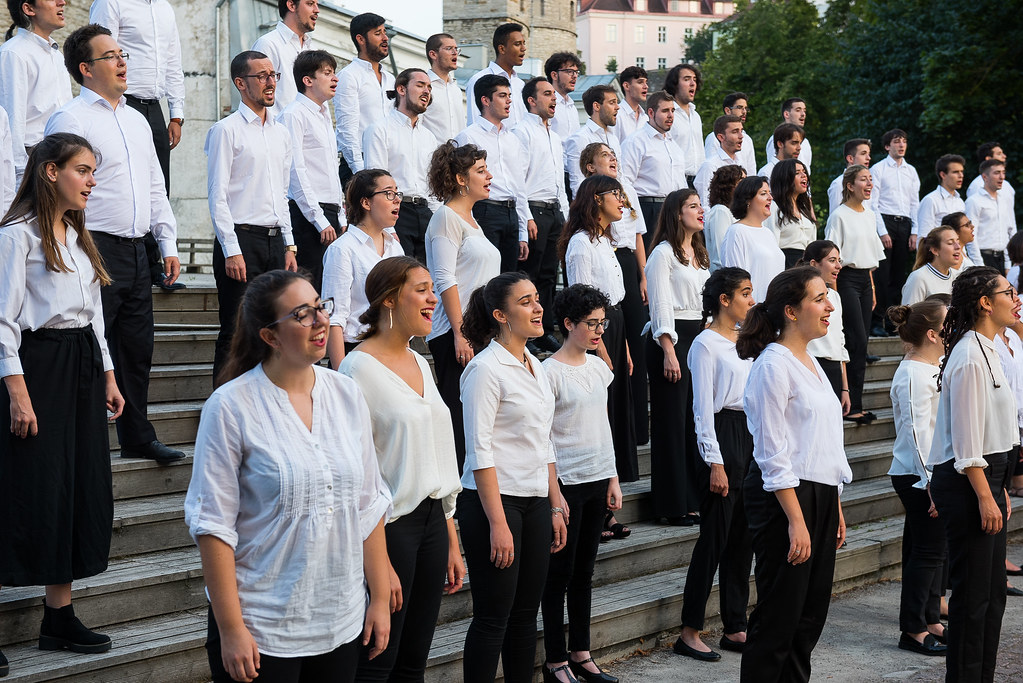 Daudzveidīgs vīriešu un sieviešu koris, ģērbies baltos kreklos un tumšās biksēs vai svārkos, stāv uz āra kāpnēm ar atvērtām mutēm, dziedot. Fonā redzamas ēkas un zaļumi.
