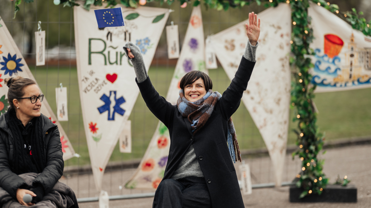 Smaidoša sieviete melnā mētelī un raibā šallē priecīgi paceļ abas rokas gaisā, kreisajā rokā turot mazu Eiropas Savienības karodziņu. Pa kreisi sēž cita sieviete. Fonā ir dekoratīvi trīsstūrveida karogi ar uzrakstiem "Rūre", "TĀ IR VESELA DZĪVE", sirds un zvaigznes simboliem, kā arī vītne ar lampiņām.