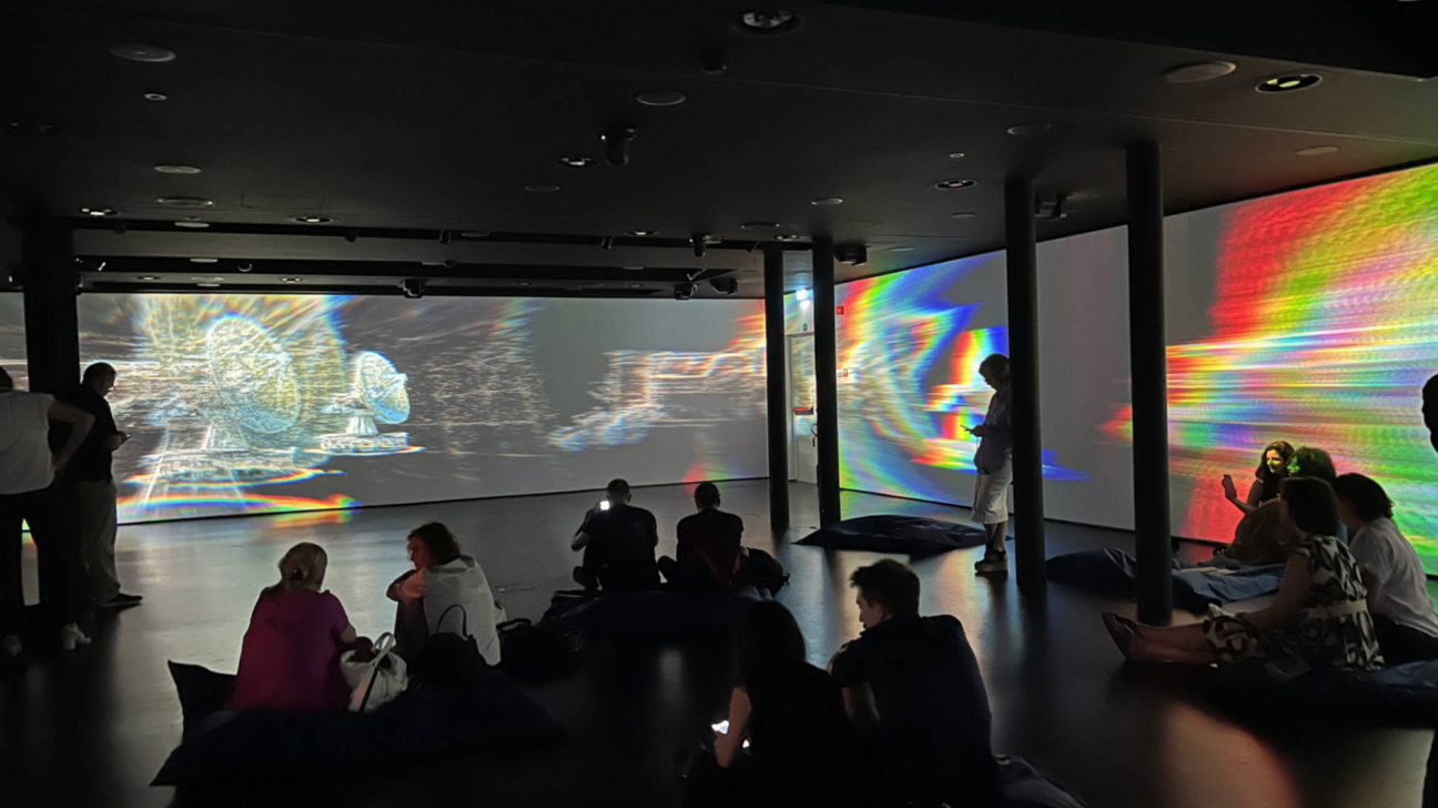 Tumšā izstāžu zālē vairāki cilvēki sēž uz sēžammaisiem vai stāv, vērojot lielas sienu projekcijas. Uz ekrāniem redzami krāsaini, abstrakti raksti, kas atgādina kļūdu attēlus, un spīdīgs satelītšķīvja attēls.