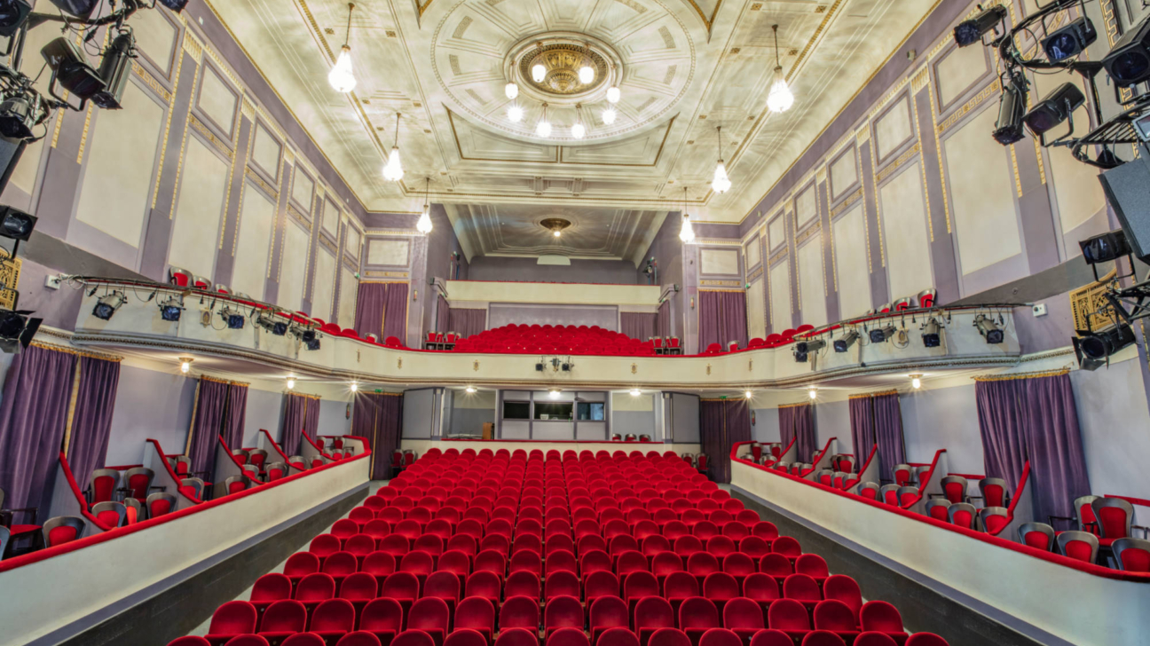 Liels, tukšs teātris vai kinoteātris ar simtiem sarkanu samta krēslu galvenajā zālē un balkonos. Telpai ir grezni rotātas gaiši violetas un zeltītas sienas, kā arī ornamentāli griesti ar lustru. Skatuve ir redzama fonā, un sānos ir redzams apgaismojuma aprīkojums.
