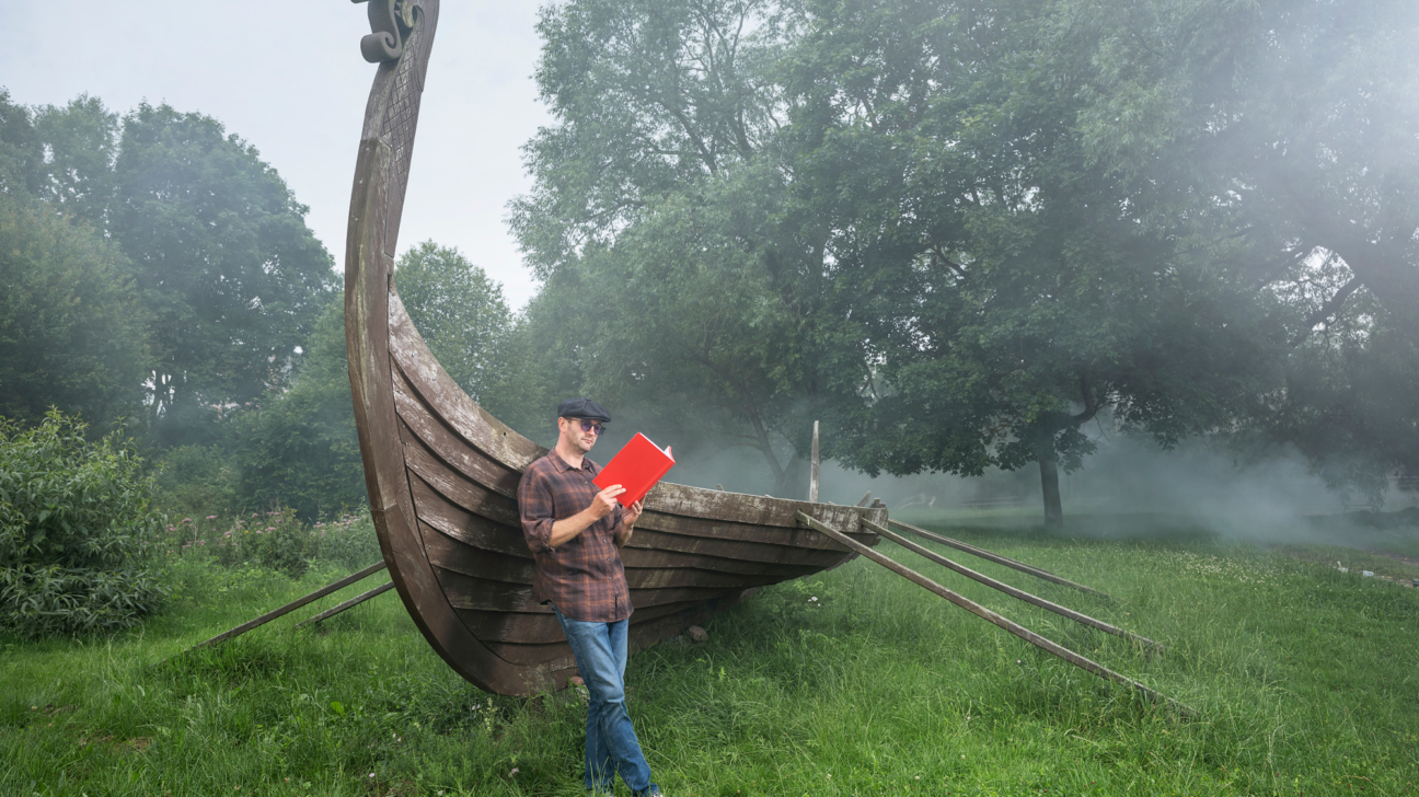 Vīrietis brūnā rūtainā kreklā, džinsos un keponā lasa sarkanu grāmatu, stāvot pie liela koka vikingu kuģa ar cirstu pūķa galvu priekšgalā, zaļā pļavā ar kokiem un dūmaku fonā.