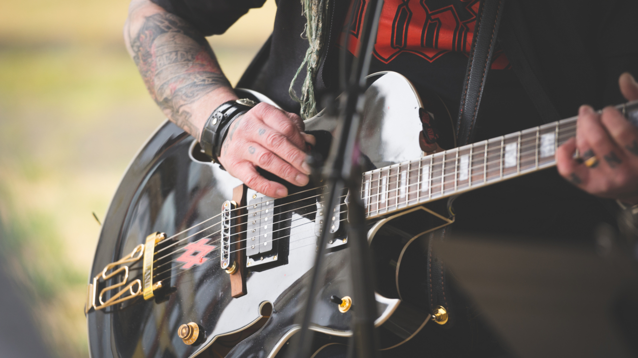 Persona ar tetovētām rokām spēlē melnu elektrisko ģitāru ar zelta detaļām. Viņa valkā melnu kreklu.