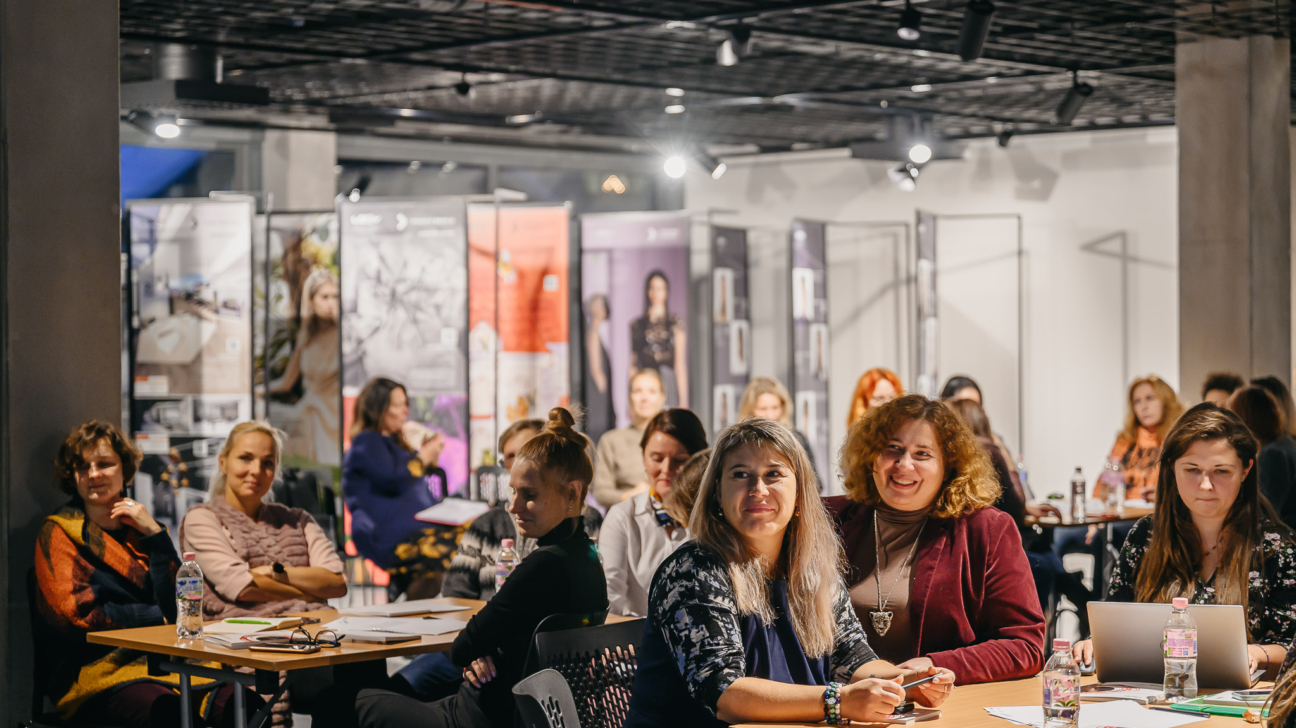 Sieviešu grupa sēž pie galdiem, apmeklējot iekštelpu pasākumu. Daudzas smaida un klausās, uz galdiem ir piezīmju grāmatiņas un ūdens pudeles, viena sieviete izmanto portatīvo datoru. Fonā ir lieli vertikāli baneri ar sieviešu attēliem.