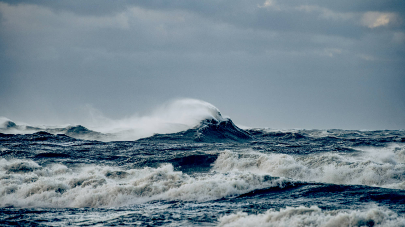 Vētraina jūra ar lieliem, tumši ziliem viļņiem, kas plīst baltās putās zem drūmām, apmākušām debesīm.