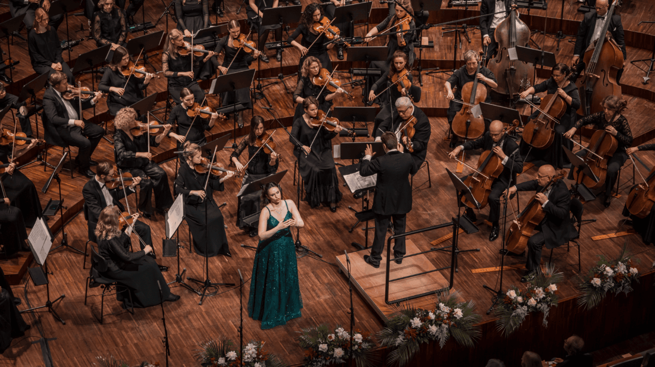 Dziedātāja mirdzoši zaļā kleitā uzstājas uz koka skatuves ar simfonisko orķestri, kura diriģents stāv uz paaugstinājuma. Mūziķi spēlē dažādus stīgu instrumentus. Skatuves priekšā ir ziedu rotājumi, un skats ir no augšas.