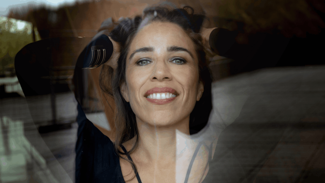 Smaidīga sieviete ar brūniem matiem un zilām acīm skatās kamerā, rokas aiz galvas. Attēlā redzama dubultā ekspozīcija ar izplūdušu āra ainavu.