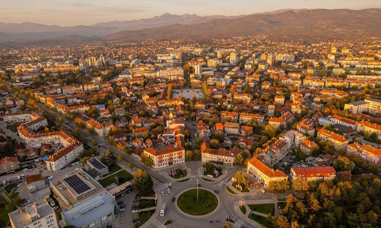 Pilsētas ainavas aerofotogrāfija saulrietā, kurā redzamas daudzas ēkas ar oranžiem un sarkaniem jumtiem, ielas, plašs apļveida krustojums un koki ar rudens lapām. Fonā redzami kalni.