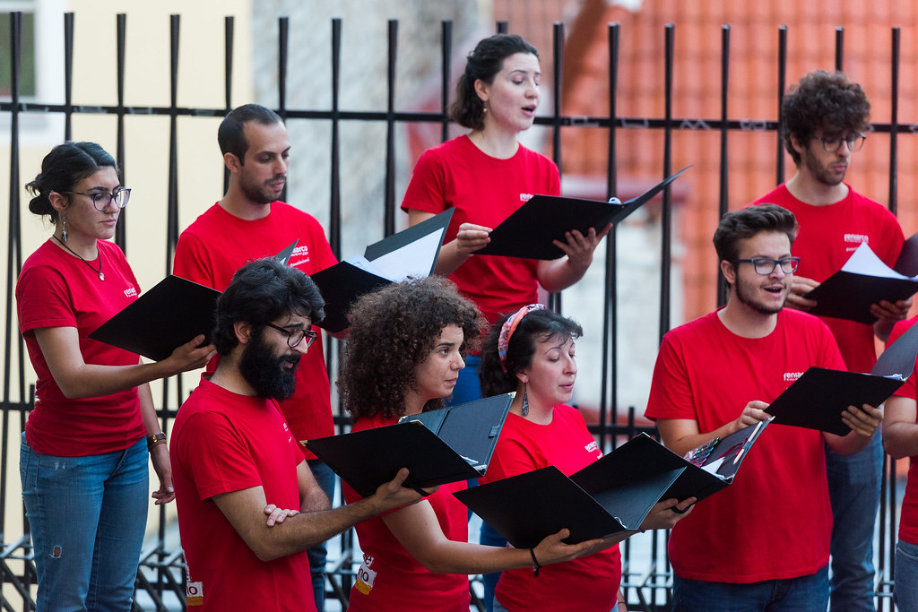 Koris dzied ārā. Dziedātāji ir tērpušies sarkanos t-kreklos ar uzrakstu "renascimento" un tur notis melnās mapēs.