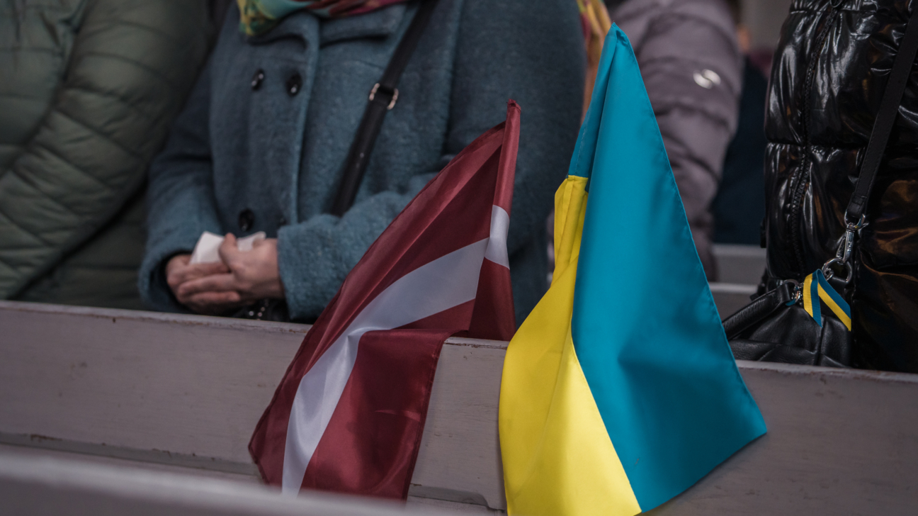 Latvijas un Ukrainas karogi novietoti viens otram blakus uz koka margām pasākumā, fonā redzami cilvēki.