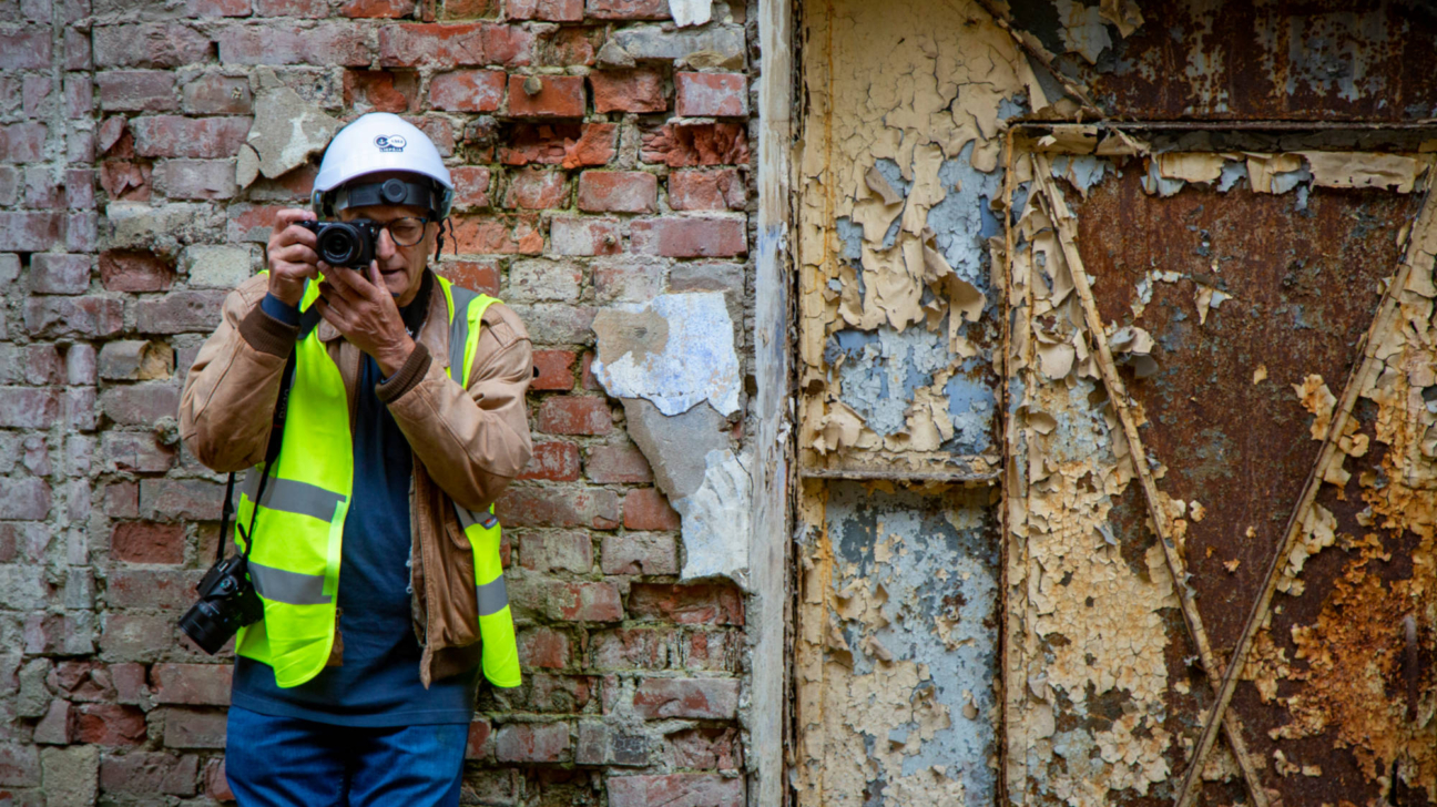 Vecāks vīrietis baltā aizsargķiverē un spilgti dzeltenā atstarojošā vestē stāv pie nolupušas ķieģeļu sienas un ar kameru rokās fotografē. Viņa labajā pusē redzamas rūsainas metāla durvis ar lobījušos krāsu.