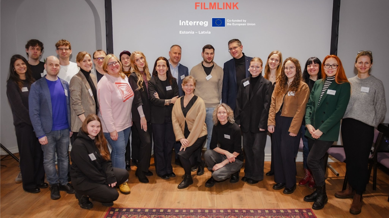 Aptuveni 25 smaidoši cilvēki, pārsvarā pieaugušie, pozē grupas fotogrāfijai iekštelpās. Viņu aizmugurē ir ekrāns ar uzrakstiem "FILMLINK", "Interreg", "Co-funded by the European Union" un "Estonia - Latvia".