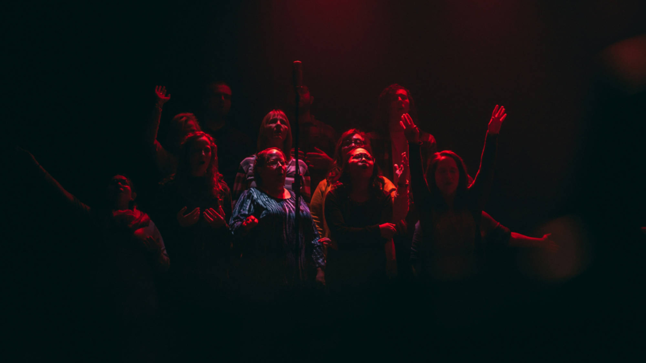 Grupa cilvēku tumšā telpā, ko izgaismo sarkana gaisma. Dažiem ir atvērtas mutes un paceltas rokas, liecinot par dziedāšanu vai pielūgsmi.
