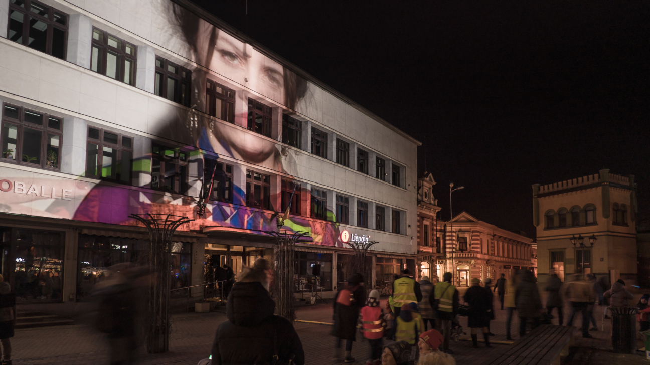 Nakts aina, kurā uz gaišas ēkas fasādes projicēta liela sievietes seja un krāsaini, abstrakti raksti. Uz ēkas redzami uzraksti "GLOBALLE" un "Liepāja". Priekšplānā un uz ielas pulcējas un staigā cilvēki, daži no tiem kustībā.