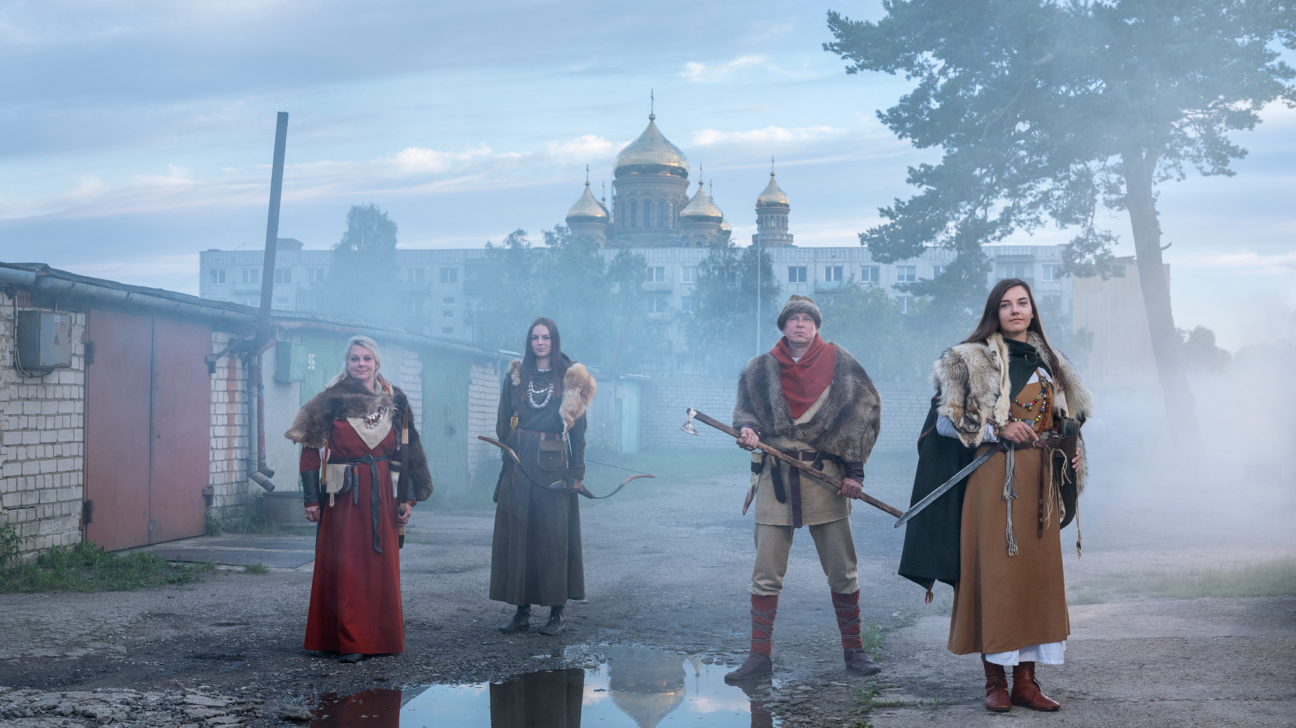 Četri cilvēki vēsturiskos tērpos ar kažokādas detaļām stāv miglainā pilsētas vidē. Kreisajā pusē ir divas sievietes, viena sarkanā kleitā, otra ar loku. Pa labi stāv vīrietis ar cirvi un sieviete ar zobenu. Fonā redzama ēka ar zelta kupoliem, daudzdzīvokļu nami, garāžas un liels koks.