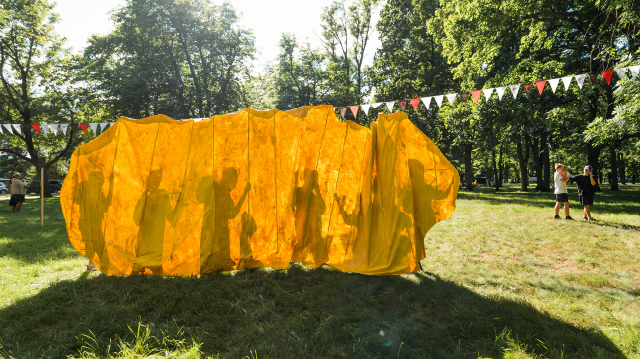 Gara, spilgti dzeltena, akordeonveidīga konstrukcija parkā, caur kuru redzami vairāku cilvēku silueti kustībā. Fonā koki un svētku karodziņu virtenes, bet labajā pusē stāv divi cilvēki.