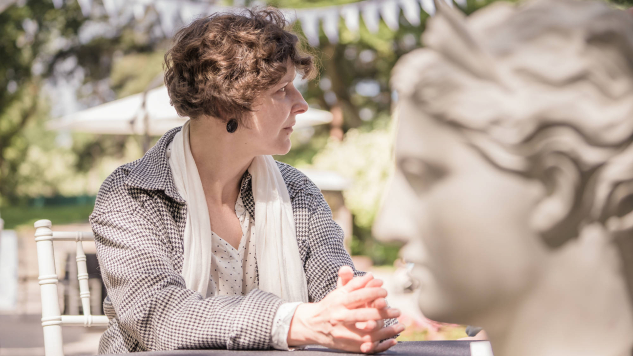 Sieviete ar tumšiem, sprogainiem matiem, ģērbusies rūtotā žaketē un baltā šallē, sēž pie galda ar saliktām rokām un skatās uz labo pusi. Priekšplānā pa labi redzams izplūdis antīkas statujas biusts, bet fonā – zaļumi un baltas dekoratīvās virtenes.