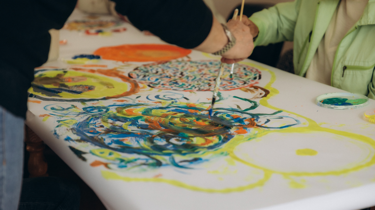 Divi cilvēki glezno ar krāsainām krāsām uz liela balta papīra, kas klāj galdu. Redzama persona melnā kreklā, kura ar otu palīdz gleznot otrai personai gaiši zaļā jakā, veidojot abstraktus rakstus.